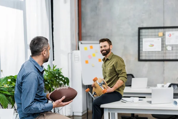 Lächelnde Geschäftsleute, die sich anschauen und American Football Ball und Longboard im Büro halten — Stockfoto