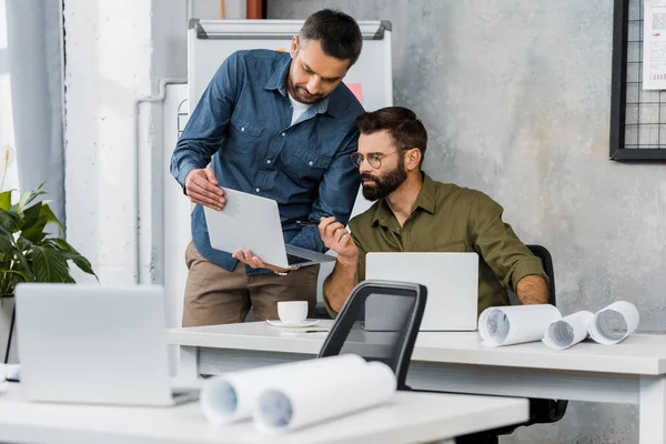 Zwei gutaussehende Geschäftsleute blicken auf Laptop im Büro, Entwürfe auf Tischplatten — Stockfoto
