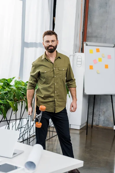 Handsome bearded businessman standing with longboard and looking at camera in office — Stock Photo