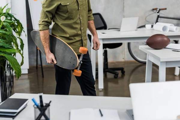 Imagen recortada de hombre de negocios de pie con longboard en la oficina - foto de stock