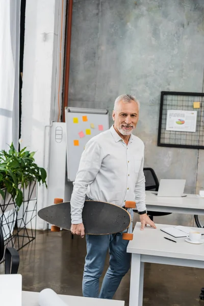 Smiling handsome middle aged businessman standing with longboard in office — Stock Photo