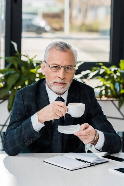 Guapo hombre de negocios de mediana edad sosteniendo taza y plato y mirando a la cámara en la oficina - foto de stock