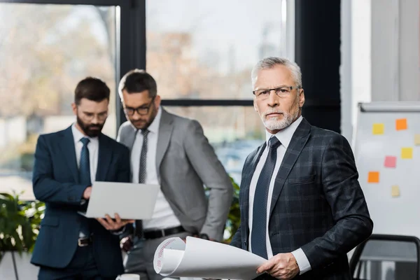 Hombres de negocios guapos en trajes y gafas de pie con portátil y planos en la oficina - foto de stock