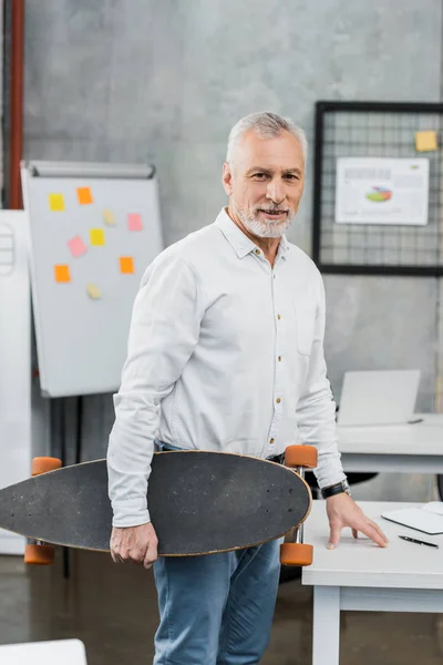 Handsome middle aged businessman holding skateboard and looking at camera in office — Stock Photo