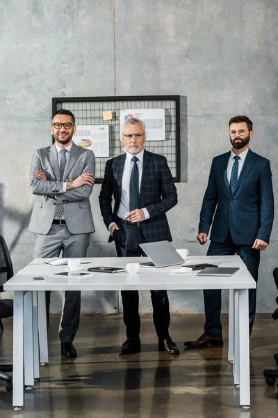 Homens de negócios bonitos em ternos de pé e olhando para a câmera no escritório — Fotografia de Stock