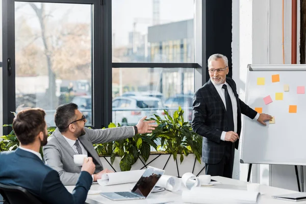 Sonriente guapo maduro hombre de negocios apuntando en flipchart en oficina - foto de stock