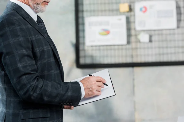 Abgeschnittenes Bild eines Geschäftsmannes mittleren Alters im Anzug, der im Büro etwas ans Notizbuch schreibt — Stockfoto