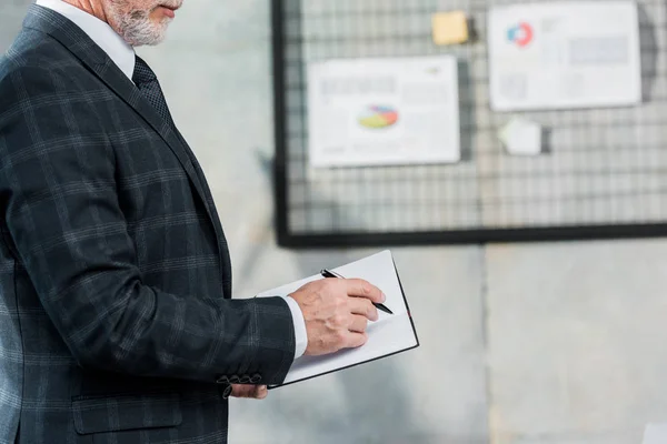 Cropped image of middle aged businessman in suit writing something to notebook in office — Stock Photo