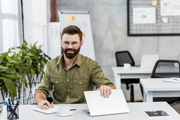 Lächelnder gutaussehender Geschäftsmann, der mit Laptop und Notizbuch am Tisch sitzt und im Büro in die Kamera schaut — Stockfoto