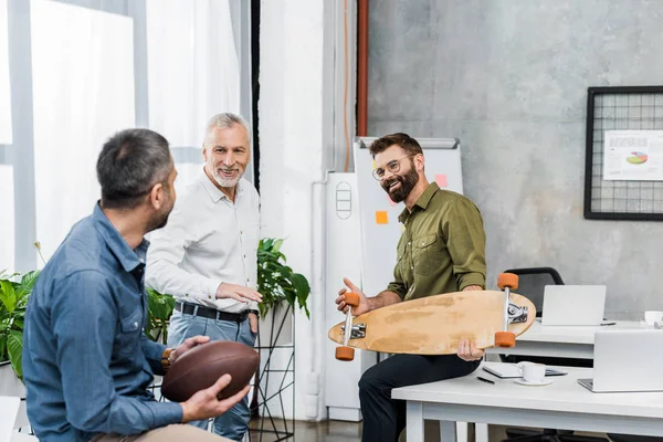Bei uomini d'affari parlando e tenendo skateboard e palla da calcio americano in ufficio — Foto stock