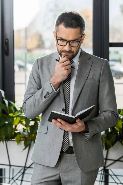 Красивый задумчивый бизнесмен в костюме и очках, глядя на блокнот в офисе — стоковое фото