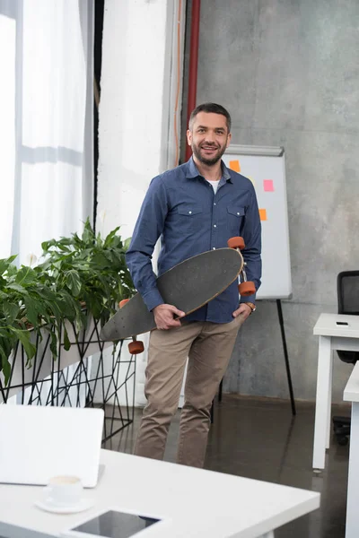 Smiling handsome businessman holding longboard and looking at camera in office — Stock Photo