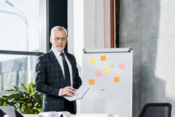 Bel homme d'affaires d'âge moyen debout près de tableau à feuilles mobiles et regardant la caméra dans le bureau — Photo de stock