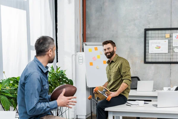 Smiling businessmen looking at each other and holding american football ball and longboard in office — Stock Photo