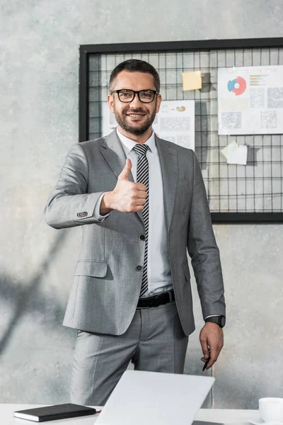 Handsome businessman showing thumb up and looking at camera in office — Stock Photo