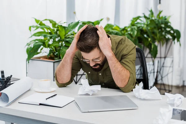 Втомлений красивий бізнесмен торкається голови і сидить за столом зі збитим папером в офісі — стокове фото