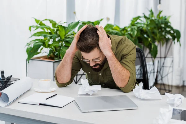 Hombre de negocios guapo cansado tocando la cabeza y sentado en la mesa con papel arrugado en la oficina - foto de stock