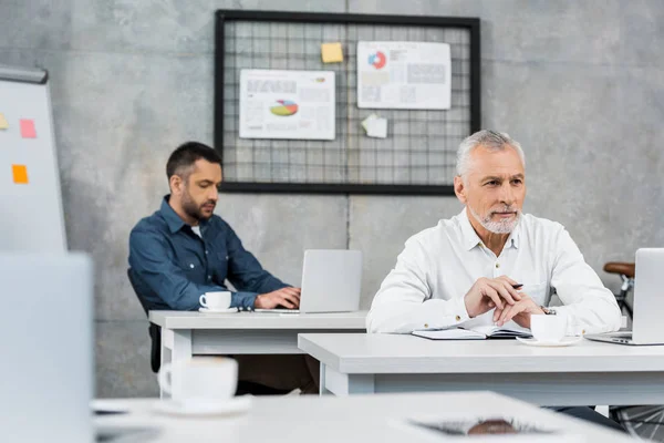 Enfoque selectivo de hombres de negocios guapos pensativos sentados en la mesa y mirando hacia otro lado en la oficina - foto de stock