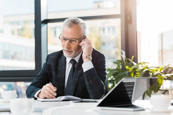 Gutaussehender Geschäftsmann mittleren Alters sitzt im Büro am Tisch und unterhält sich per Smartphone — Stockfoto