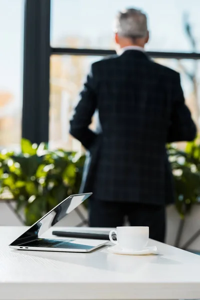 Вид ззаду бізнесмена середнього віку, що стоїть в офісі, ноутбуці і чашці чаю на столі — стокове фото