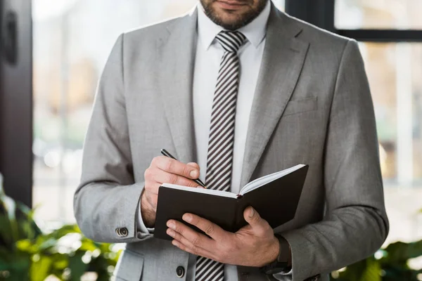 Обрезанный образ бизнесмена в костюме, пишущего что-то в блокнот в офисе — стоковое фото