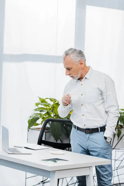 Красивий бізнесмен середнього віку тримає чашку чаю і дивиться на ноутбук в офісі — стокове фото
