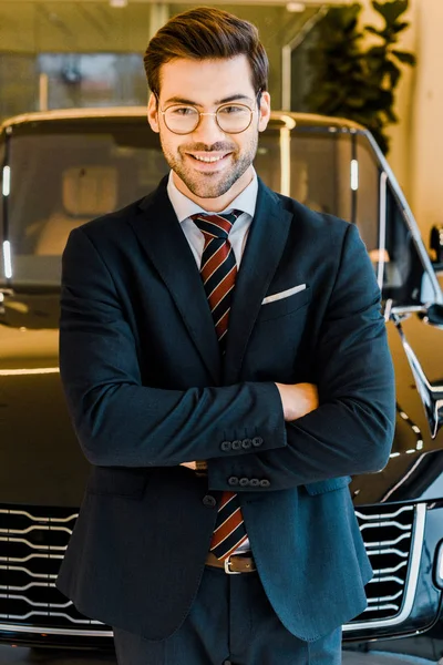 Laughing businessman in eyeglasses posing with crossed arms near black automobile — Stock Photo