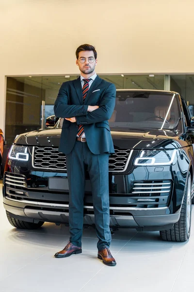 Low angle view of businessman in eyeglasses posing with crossed arms near black automobile — Stock Photo