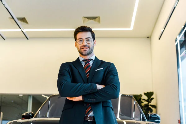 Low angle view of smiling businessman in eyeglasses posing with crossed arms near black automobile — Stock Photo