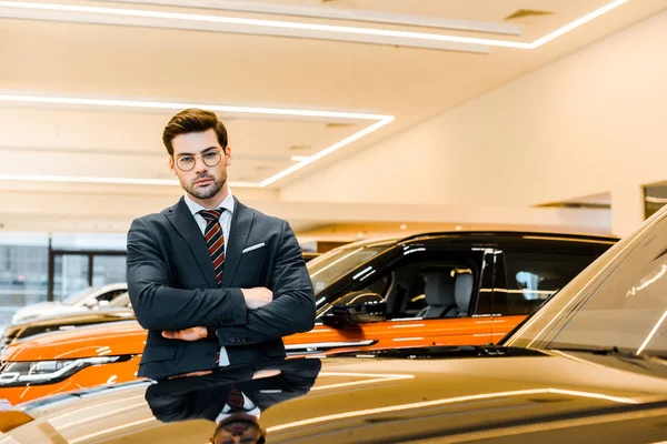 Confident businessman in eyeglasses posing with crossed arms near black automobile — Stock Photo