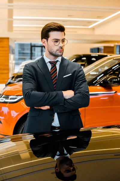 Serio hombre de negocios en gafas posando con los brazos cruzados cerca de automóvil negro - foto de stock