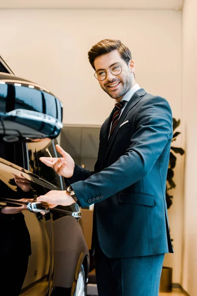 Cheerful male car dealer in eyeglasses pointing at black automobile in car salon — Stock Photo