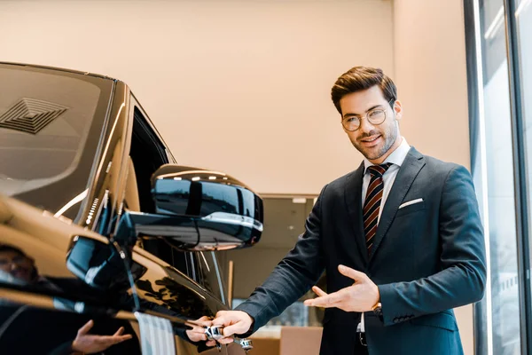 Smiling male car dealer in eyeglasses pointing at black automobile in car salon — Stock Photo