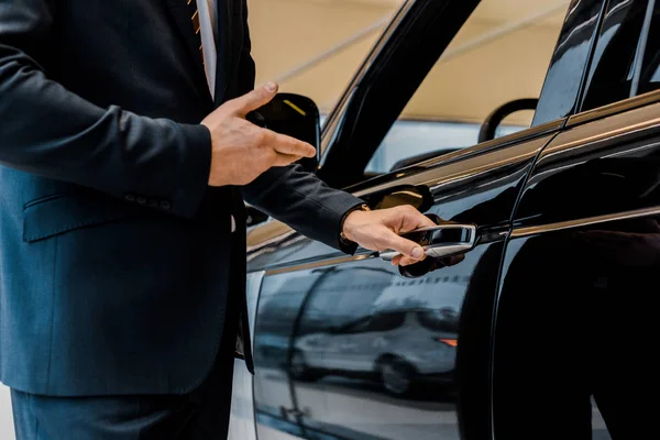 Cropped image of male car dealer opening door and pointing at black automobile in car salon — Stock Photo