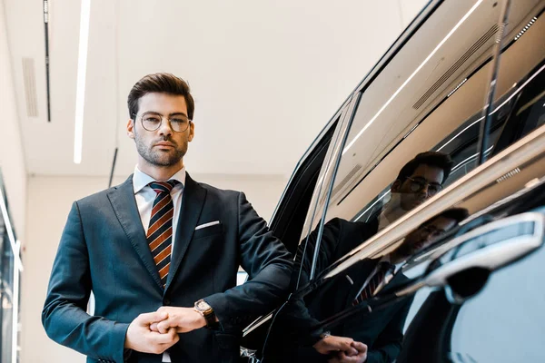 Low angle view of businessman in eyeglasses posing near automobile in car salon — Stock Photo