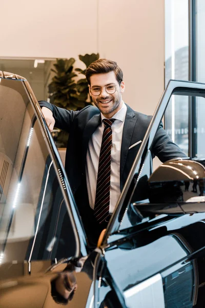 Laughing businessman in eyeglasses posing near automobile in car salon — Stock Photo