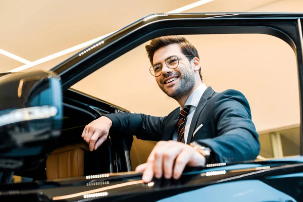 Vista de ángulo bajo de hombre de negocios feliz en anteojos posando cerca de automóvil en salón de coches - foto de stock