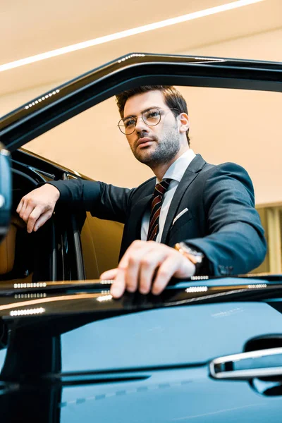 Low angle view of confident businessman in eyeglasses posing near automobile in car salon — Stock Photo