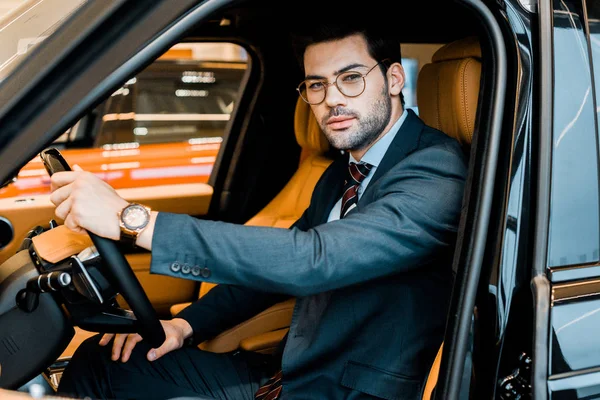 Stylish businessman in eyeglasses looking at camera while sitting in luxury car — Stock Photo