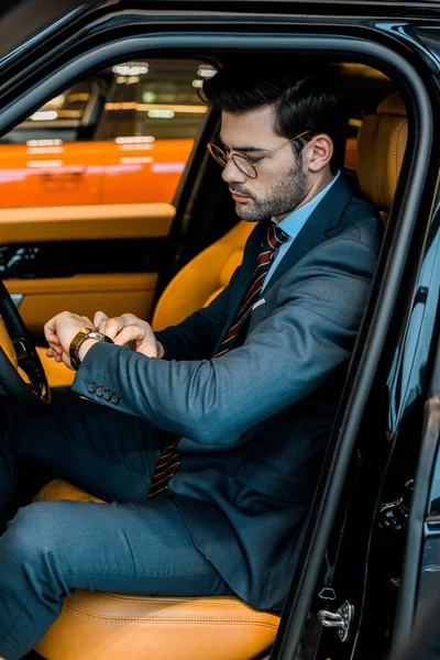 Handsome businessman in eyeglasses checking watch in luxury car — Stock Photo
