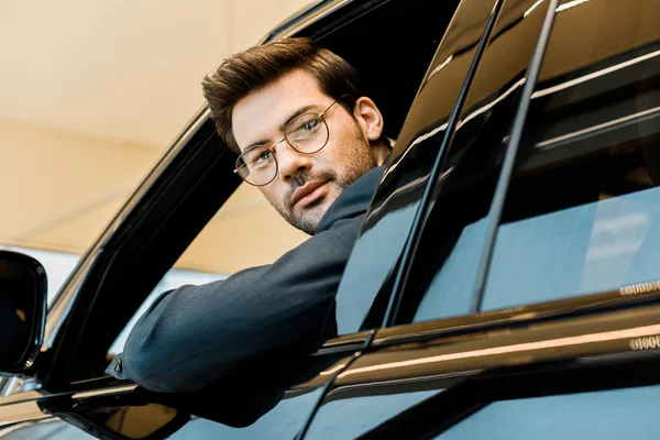 Selective focus of stylish businessman in eyeglasses looking out from window of car — Stock Photo