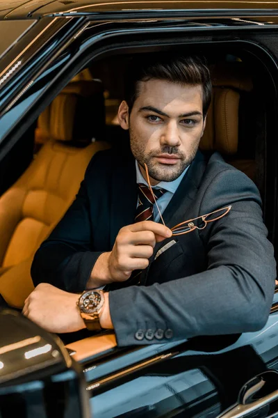 Guapo hombre de negocios con reloj de lujo sosteniendo gafas mientras está sentado en el coche - foto de stock
