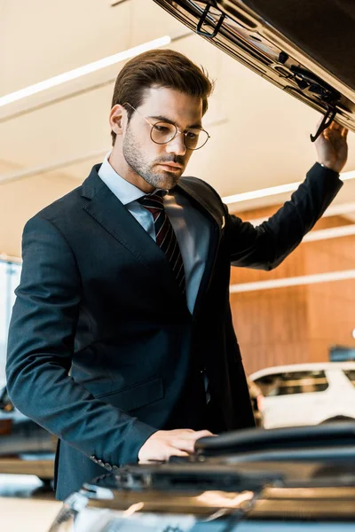 Low angle view of businessman in eyeglasses looking inside opened car hood of black automobile — Stock Photo