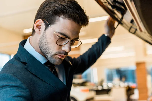 Close up view of businessman in eyeglasses looking inside opened car hood of black automobile — Stock Photo