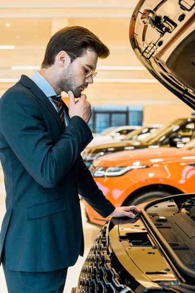 Seitenansicht eines nachdenklichen Geschäftsmannes, der auf die geöffnete Motorhaube eines schwarzen Autos blickt — Stockfoto