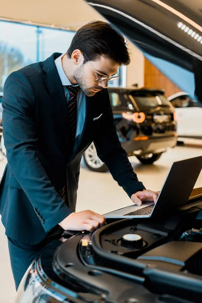 Focado jovem empresário em óculos verificando capuz de carro com laptop — Fotografia de Stock