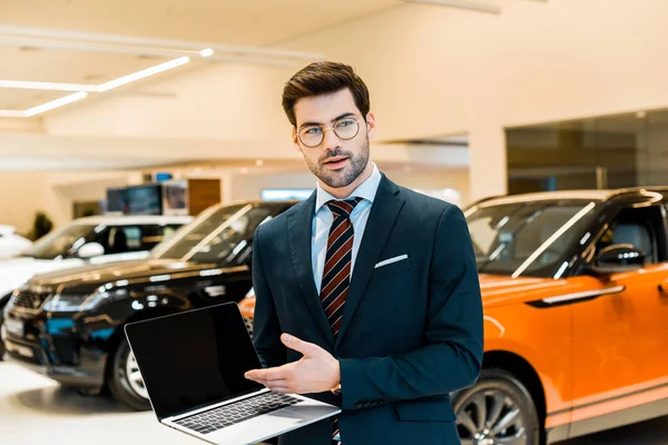 Smiling male car dealer in eyeglasses pointing at laptop with blank screen in car salon — Stock Photo