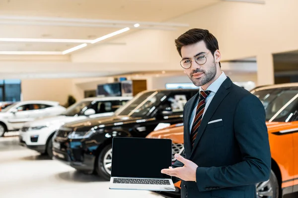 Glücklicher männlicher Autohändler mit Brille zeigt auf Laptop mit leerem Bildschirm im Autosalon — Stockfoto