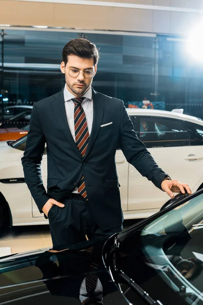 Handsome businessman in eyeglasses choosing car in dealership salon — Stock Photo