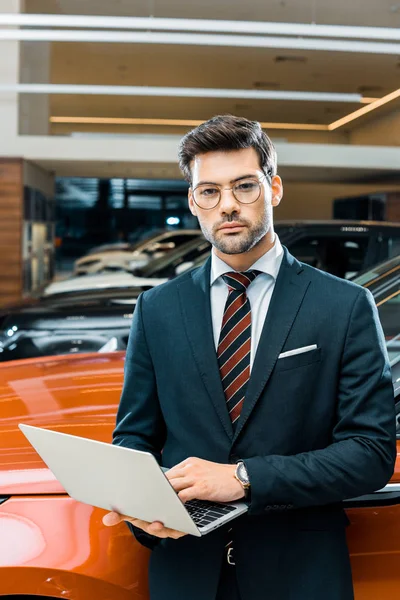 Stylish young businessman in eyeglasses using laptop near automobile — Stock Photo